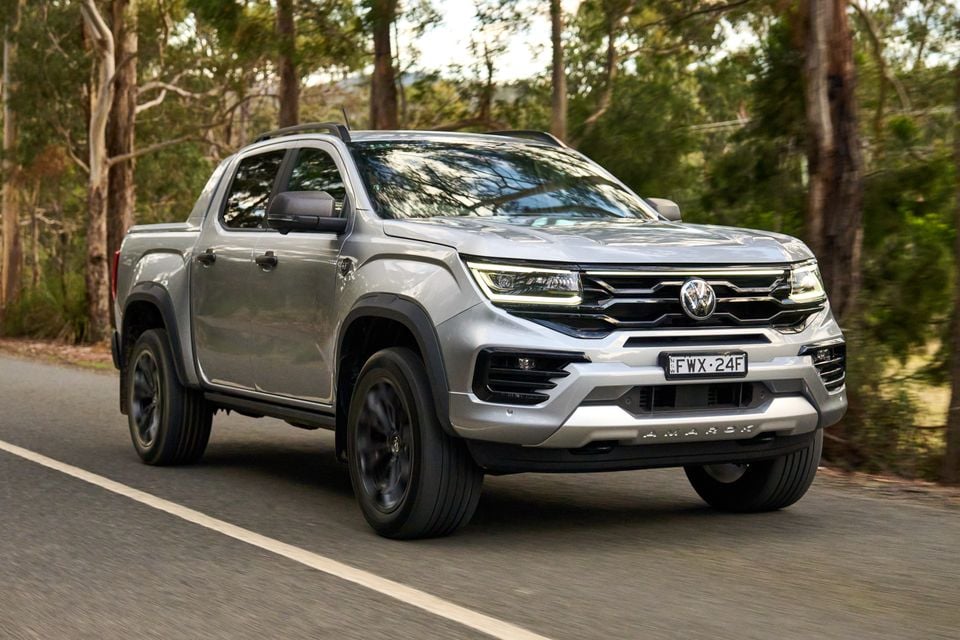 A silver Volkswagen Amarok dual-cab ute driving on a tree-lined road, seen from the front three-quarter angle