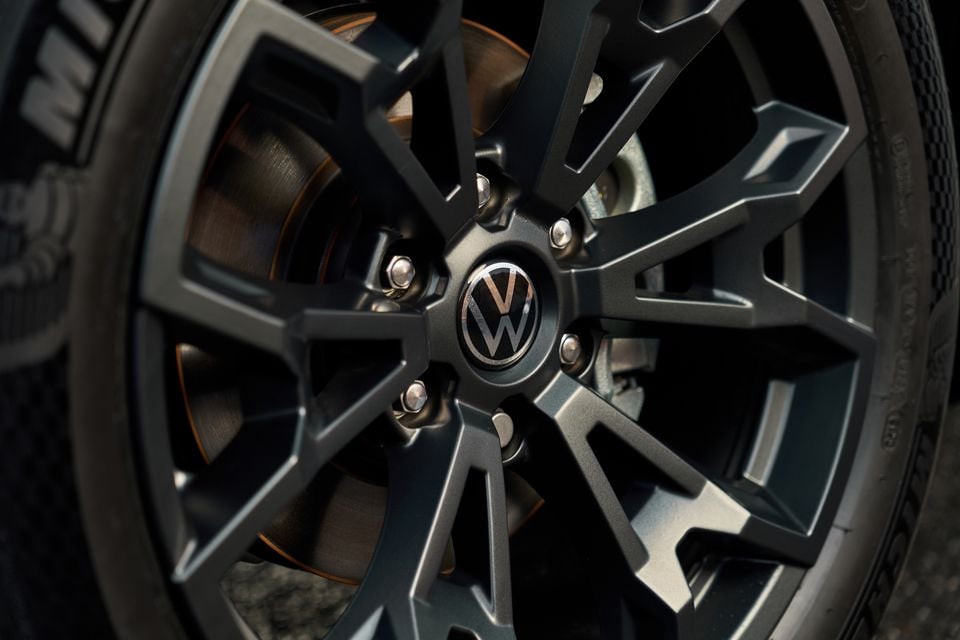 Close-up of a Volkswagen alloy wheel with a dark multi-spoke design, centre cap bearing the VW logo, and orange-accented brake caliper visible behind the rotor.