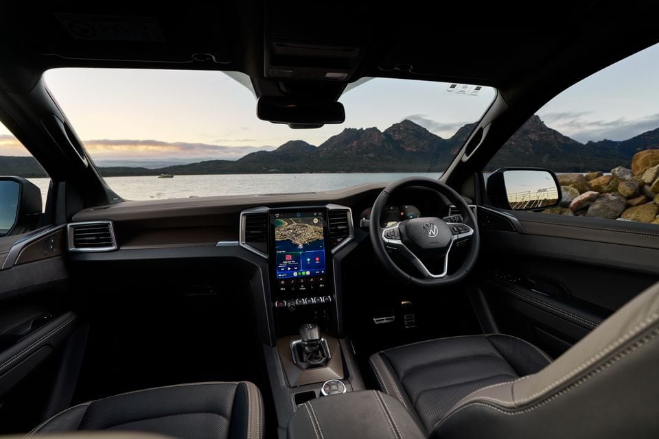 Interior of a Volkswagen Amarok showing the dashboard, large vertical touchscreen infotainment display, steering wheel, and front seats, with a scenic mountain and waterway view through the windscreen