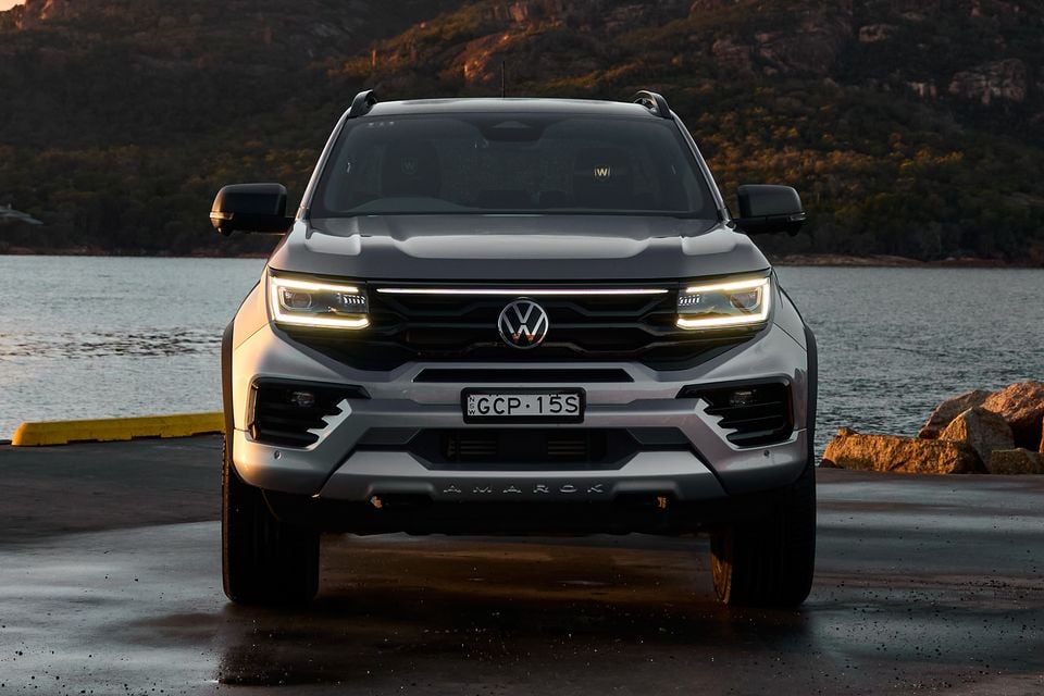 A silver Volkswagen Amarok ute seen from the front, with headlights on, parked near a waterfront at dusk with rocky hills in the background.