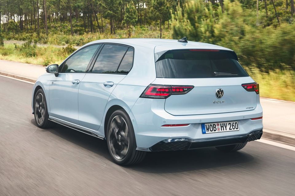 A light blue Volkswagen Golf hybrid hatchback seen from the rear three-quarter angle while driving on a road lined with trees