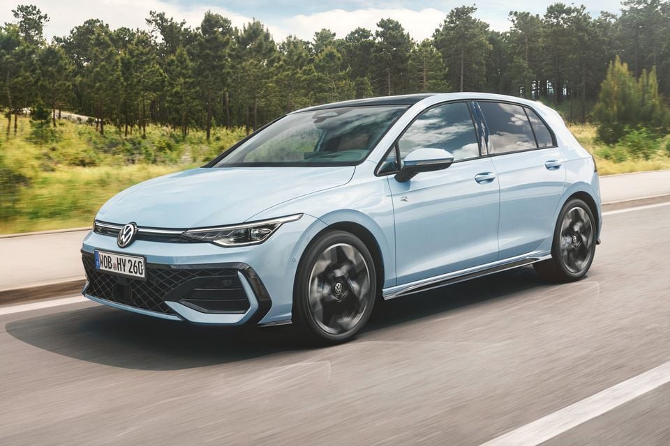 A light blue Volkswagen Golf R hatchback driving on a road, seen from the front-side angle, with trees in the background.