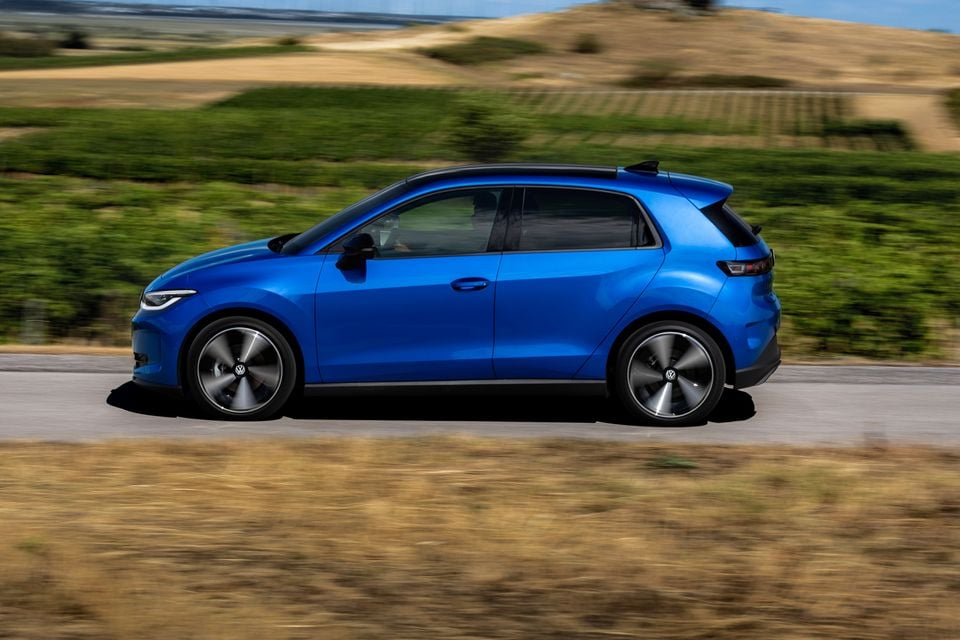 A blue Volkswagen ID. Polo hatchback seen from the side while driving on a road, with green hills and countryside in the background.