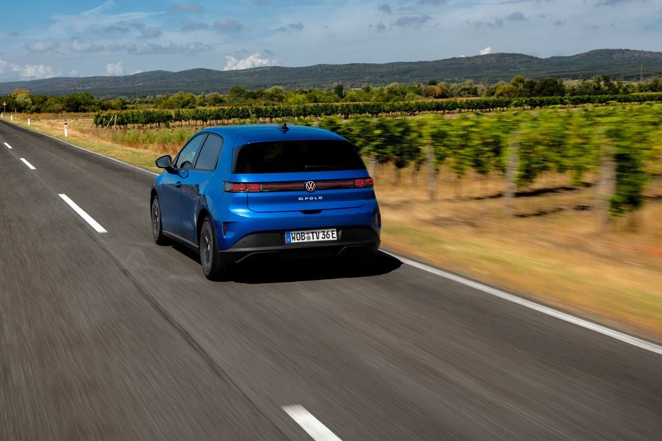 A blue Volkswagen ID. Polo hatchback seen from the rear while driving on a country road, with vineyards and hills in the background.