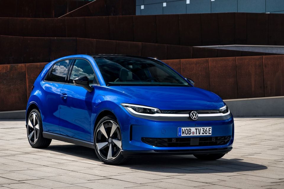 A blue Volkswagen ID. Polo hatchback seen from the front-left side, parked on a paved area in front of a rustic metal wall.