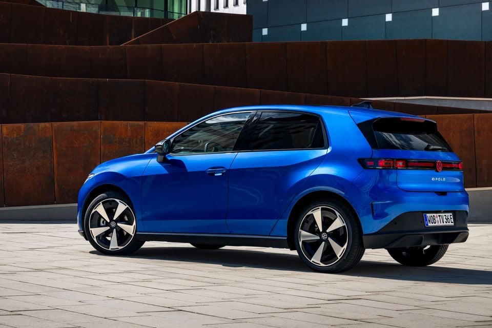 A blue 2027 Volkswagen ID. Polo hatchback seen from the rear three-quarter angle, parked on a paved surface in front of a rusted metal wall.