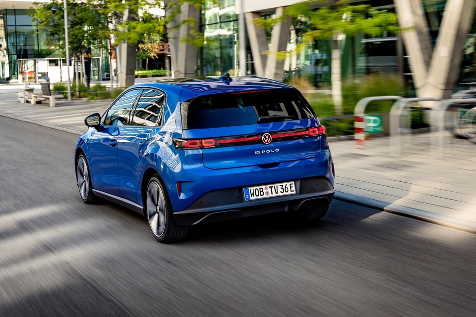 A blue 2027 Volkswagen ID. Polo hatchback seen from the rear three-quarter angle while driving through an urban street