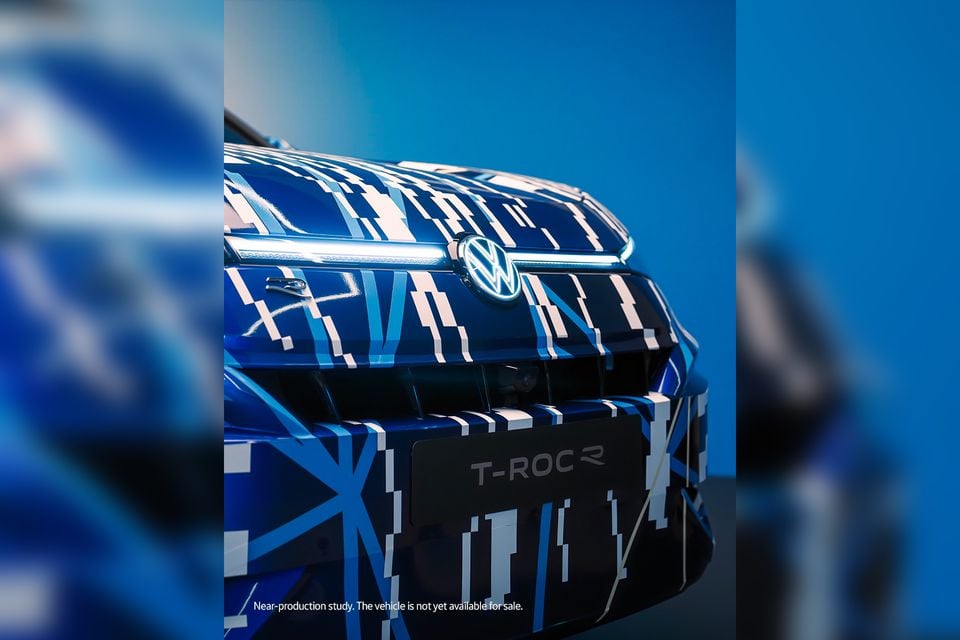Close-up of the Volkswagen T-Roc R front grille and bonnet, wrapped in blue and white camouflage, with an illuminated VW badge and T-ROC R nameplate visible
