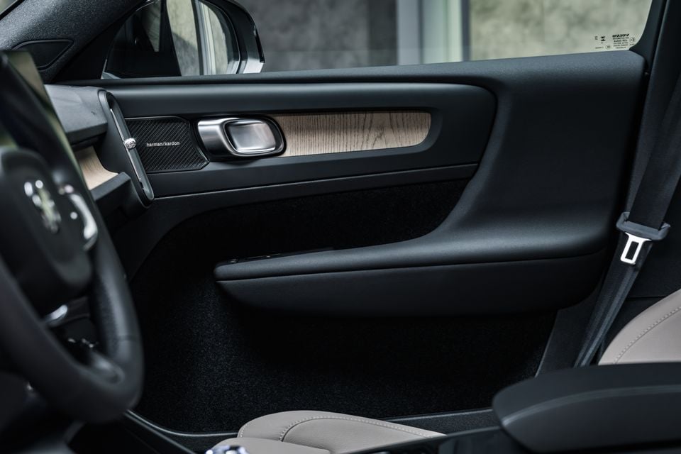 Interior door panel of a Volvo featuring a Harman Kardon speaker, wood trim inlay, and brushed metal door handle, with light tan leather seats visible in the background