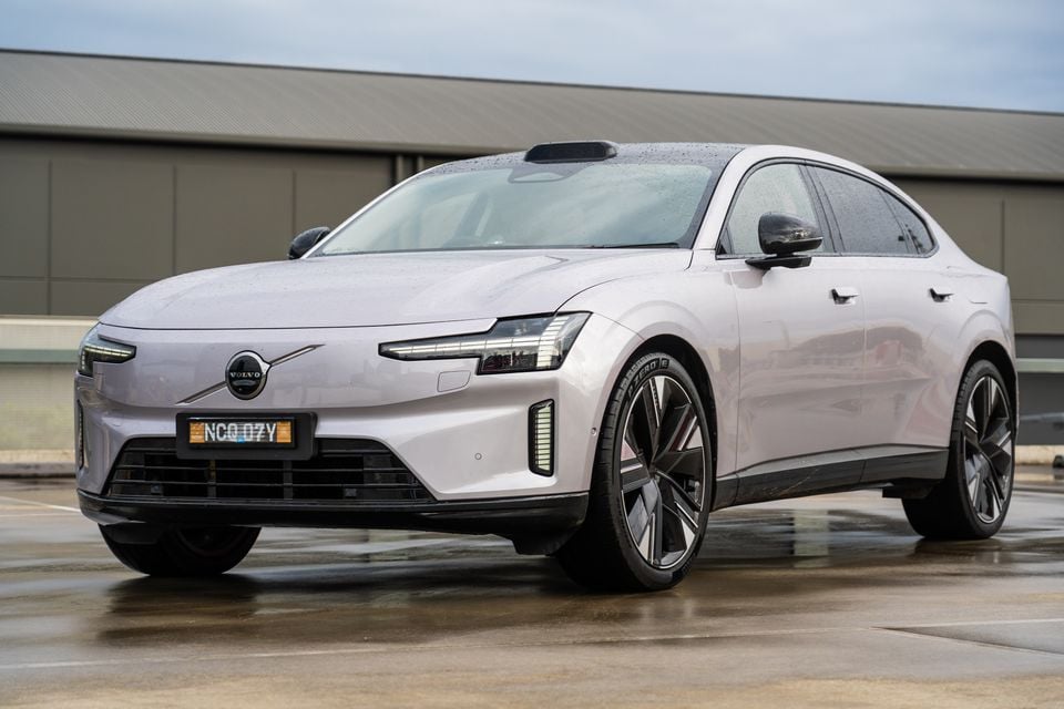 A light grey Volvo ES90 electric sedan seen from the front three-quarter angle on a wet surface near an industrial building