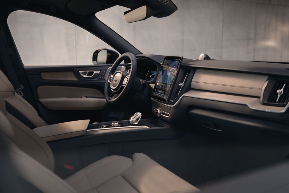 Interior of a Volvo XC60 showing the dashboard, large portrait touchscreen infotainment display, crystal gear selector, steering wheel, and light-coloured leather seats