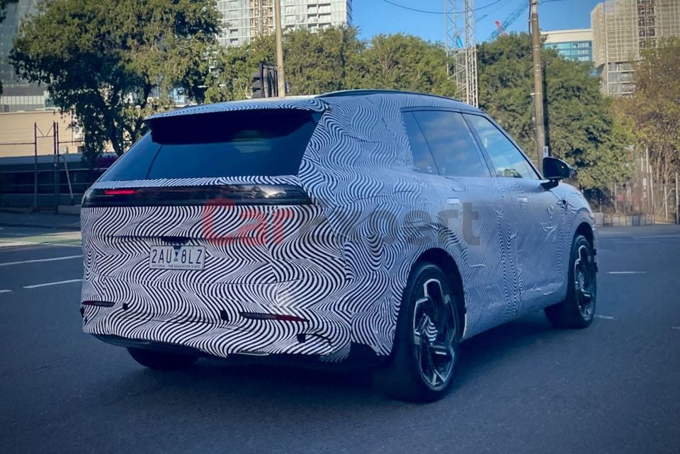 A camouflage-wrapped Xpeng G9L SUV prototype seen from the rear quarter while being tested on a city street