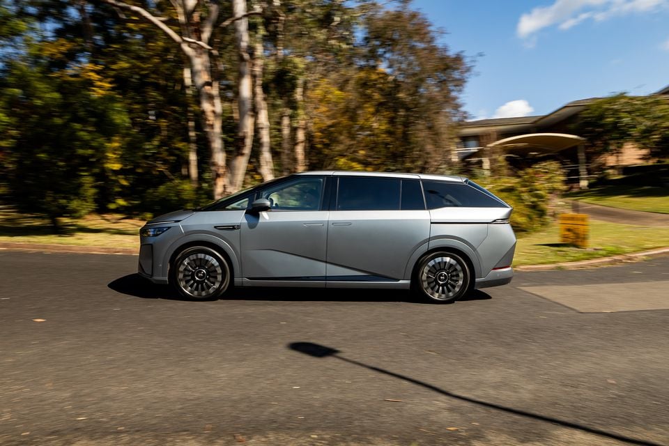 A silver 2027 Xpeng X9 large electric MPV seen from the side, driving through a suburban Australian street lined with gum trees