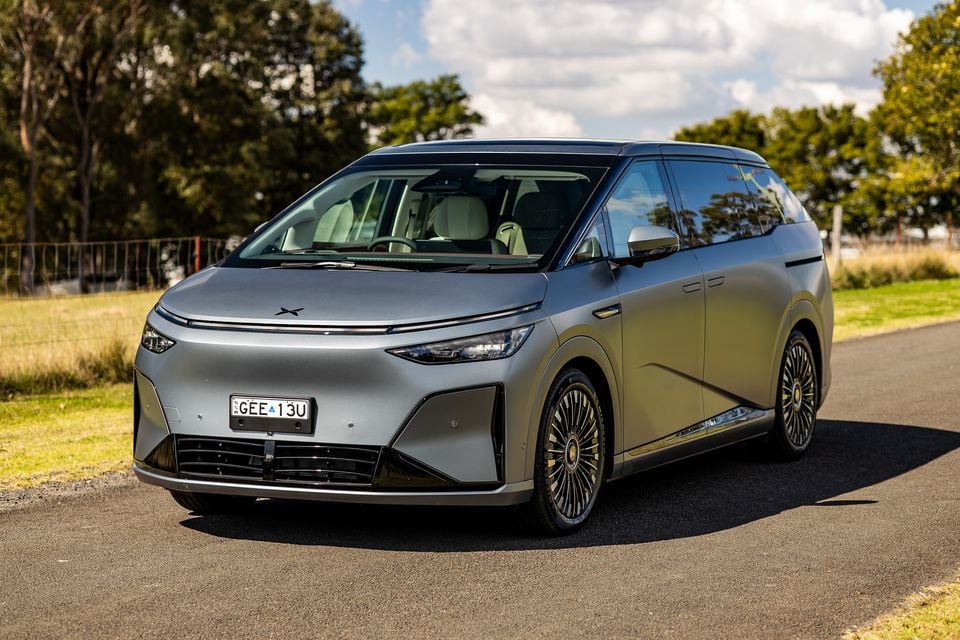 A silver 2027 Xpeng X9 electric people-mover seen from the front-side angle, parked on a road surrounded by trees and grass