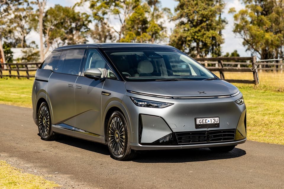 A silver 2027 Xpeng X9 electric minivan seen from the front-side angle, parked on a rural road with trees and a fence in the background.