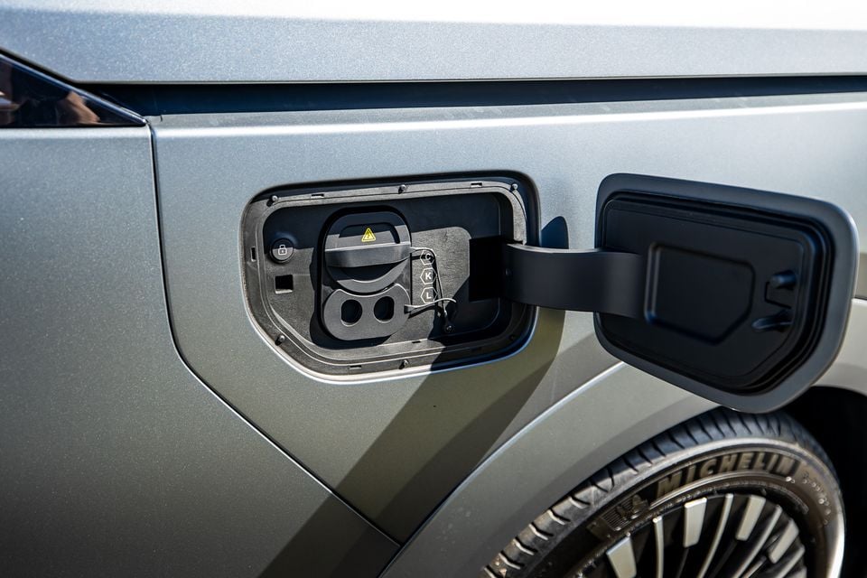 Close-up of the open charging port on a silver 2027 Xpeng X9, showing dual charge connectors with a Michelin-tyred wheel visible below