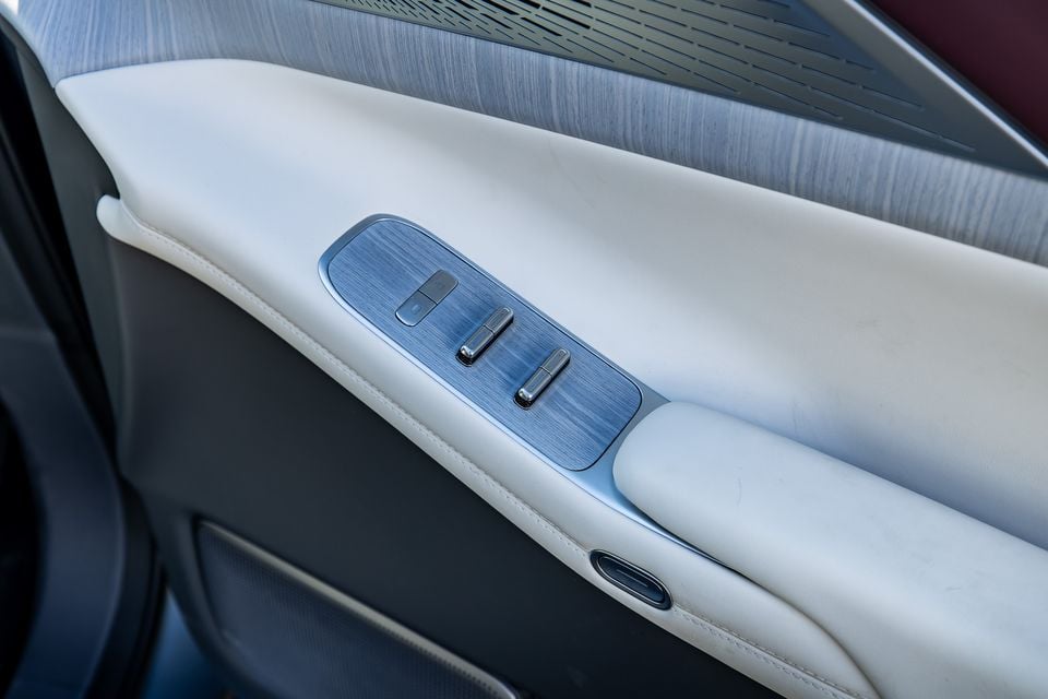 Close-up of the driver's door panel in a 2027 Xpeng X9, showing window control switches on a brushed metal panel with cream leather trim and wood-look accents.