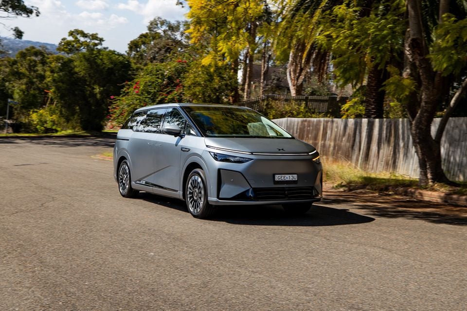 A silver 2027 Xpeng X9 electric people mover seen from the front-side angle, driving on a suburban Australian street lined with trees