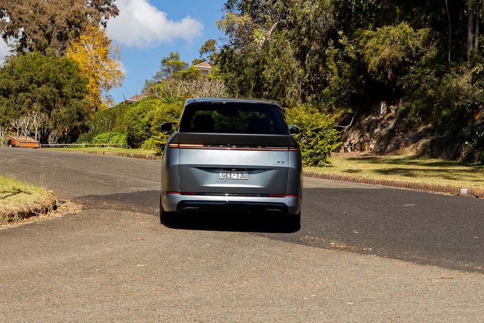 Rear view of a grey XPeng X9 large MPV parked on a suburban road, surrounded by trees and greenery