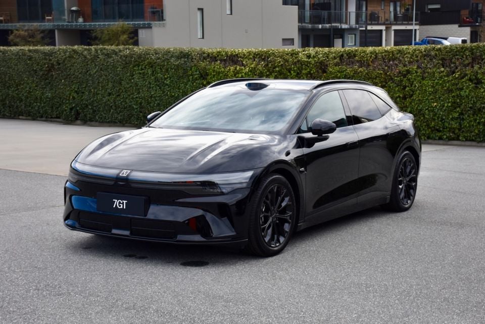 A black Zeekr 7GT seen from the front three-quarter angle, parked on a street with a hedge and apartments in the background.