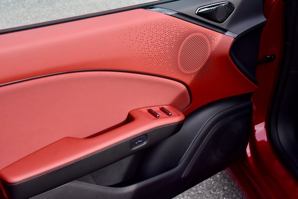 Red leather door trim panel of a car interior, showing a perforated speaker grille, window controls, and contrast stitching