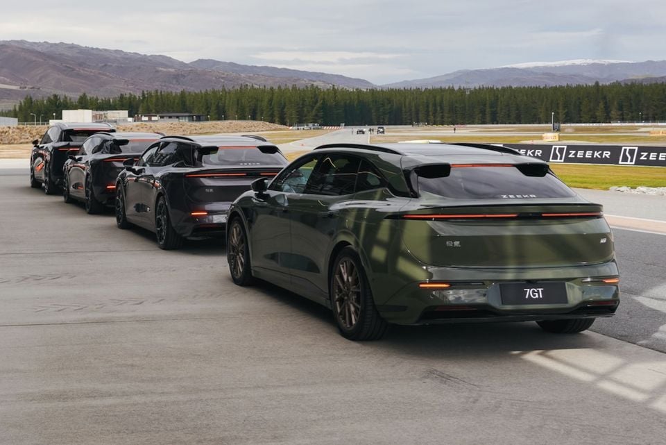 A green Zeekr 7GT and other Zeekr models lined up from the rear at a racetrack, with mountains and pine trees in the background.