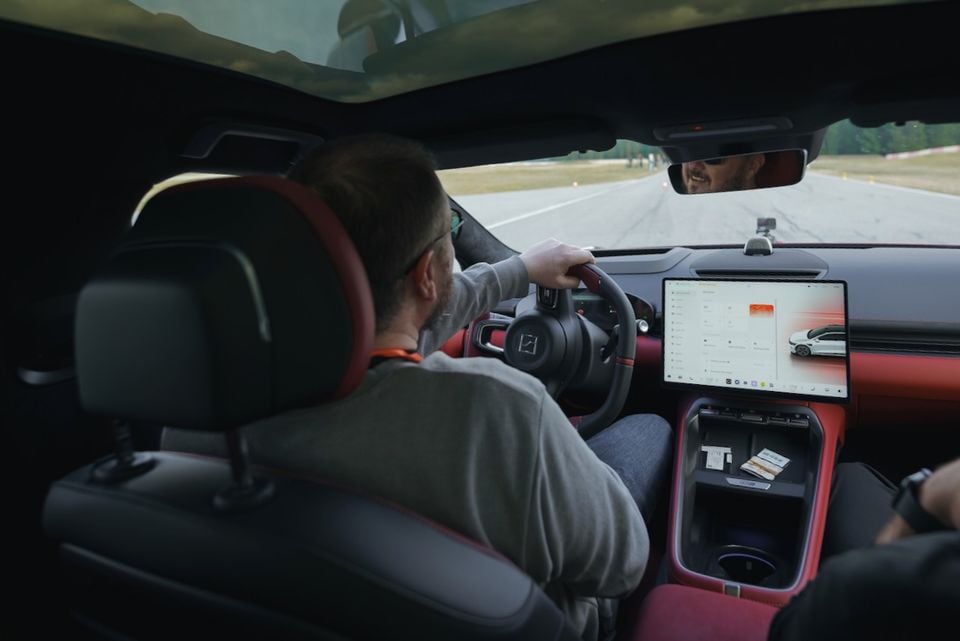 Interior view of a driver behind the wheel of a Zeekr 7GT, showing the red and black cabin, large central touchscreen infotainment display, and a track road ahead through the windscreen.