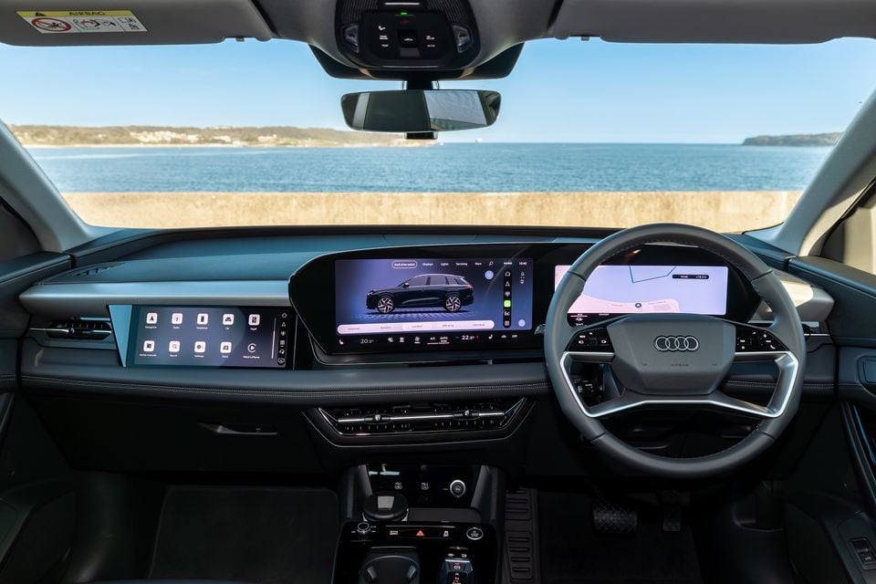 Audi Q6 e-tron interior showing the wide curved infotainment display, digital instrument cluster, and Audi steering wheel, with a coastal view through the windscreen