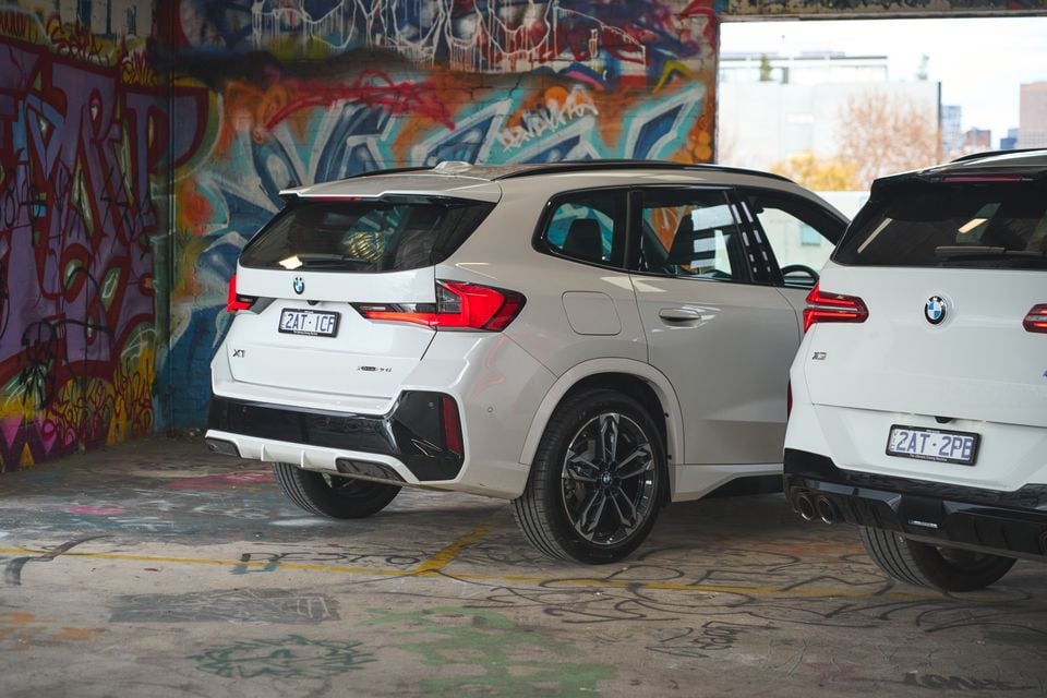 Rear three-quarter view of two white BMW SUVs — an X1 in the foreground and an X3 to the right — parked inside a graffiti-covered building in Australia.