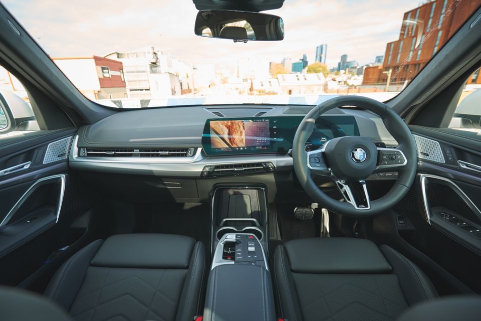 BMW SUV interior showing the dashboard, curved infotainment touchscreen, M Sport steering wheel, centre console, and front seats, with a city skyline visible through the windscreen
