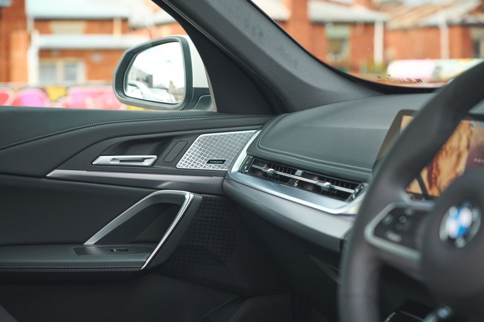 Interior view of a BMW showing the passenger door panel with a Harman Kardon speaker grille, side mirror, dashboard, air vents, and steering wheel