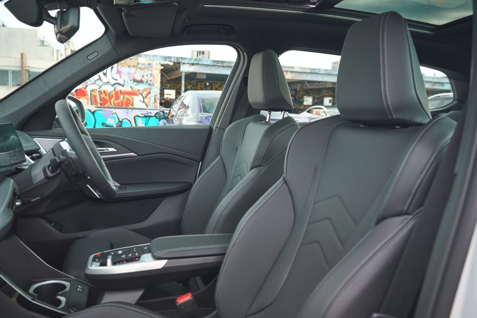 Interior view of a BMW SUV showing dark leather front seats, centre console, steering wheel, and large infotainment screen, with graffiti-covered walls visible through the windows.