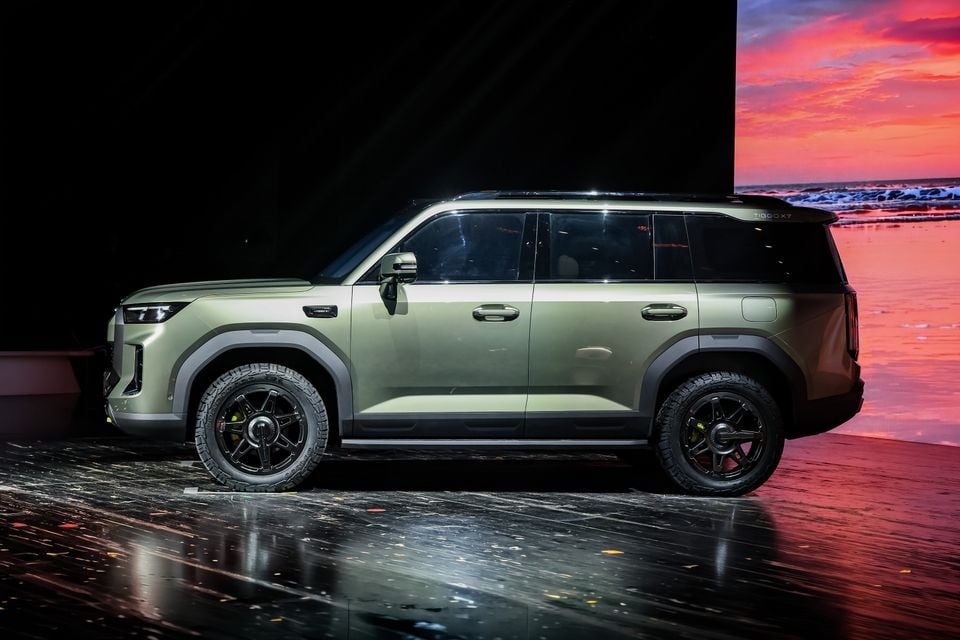 A green 2027 Chery Tiggo X7 SUV seen side-on, displayed on a stage in front of a sunset beach backdrop