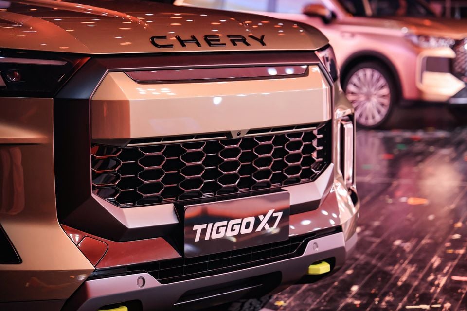 Close-up of the front grille and bonnet lettering on a bronze 2027 Chery Tiggo X7, with another Chery SUV displayed in the background