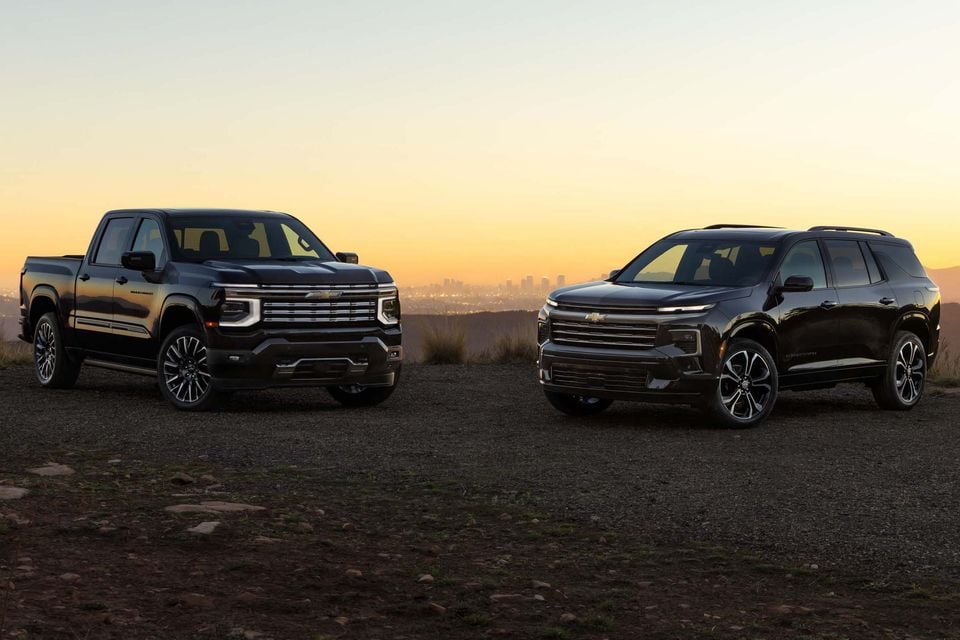 Black 2027 Chevrolet Silverado High Country pickup and Traverse High Country SUV parked on a hilltop at sunset with a city skyline behind