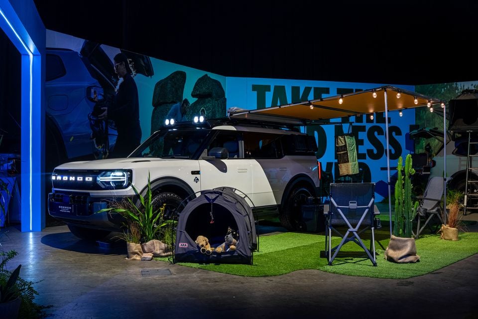 A white Ford Bronco Sport prototype on display at an indoor expo, fitted with roof lights and an ARB awning, surrounded by a camping-themed setup including a pet tent, camp chair, and cactus plants.