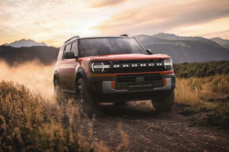 An orange Ford Bronco Basecamp SUV driving along a dusty dirt track with mountains and a dramatic sunset sky in the background
