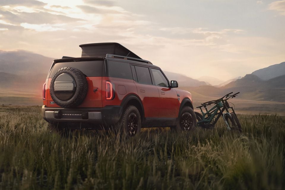 A red Ford Bronco Basecamp SUV seen from the rear three-quarter angle, with a rooftop tent popped open, parked in an open grassy field at sunset with mountains in the background and mountain bikes alongside