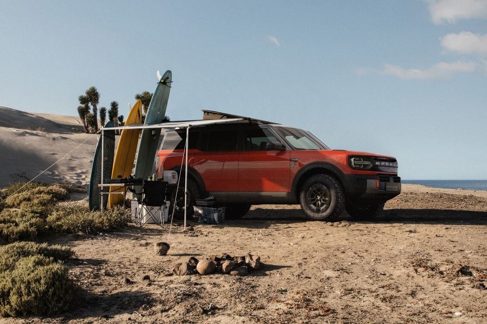 A red Ford Bronco Sport SUV set up as a beach camp with a roof awning and surfboards leaning against a rack, parked on a sandy coastal area with sand dunes and the ocean in the background.