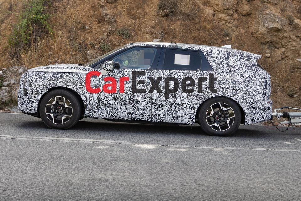 A camouflaged Kia Sportage SUV prototype seen from the side while being tested on a public road, towing a trailer