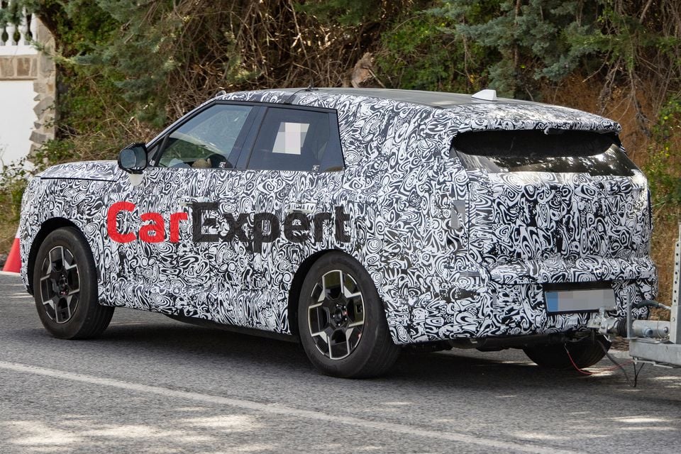 A camouflaged 2027 Kia Sportage SUV prototype seen from the rear three-quarter angle, towing a trailer during testing on a public road