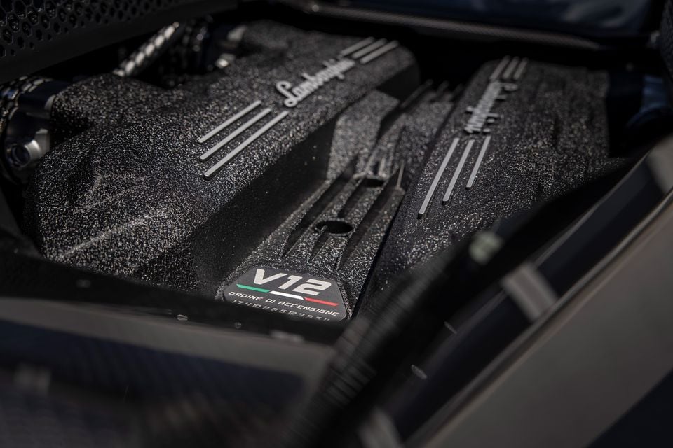 Close-up of the Lamborghini V12 engine bay, showing the textured engine covers with Lamborghini script badging and a V12 firing order plaque with Italian flag detail