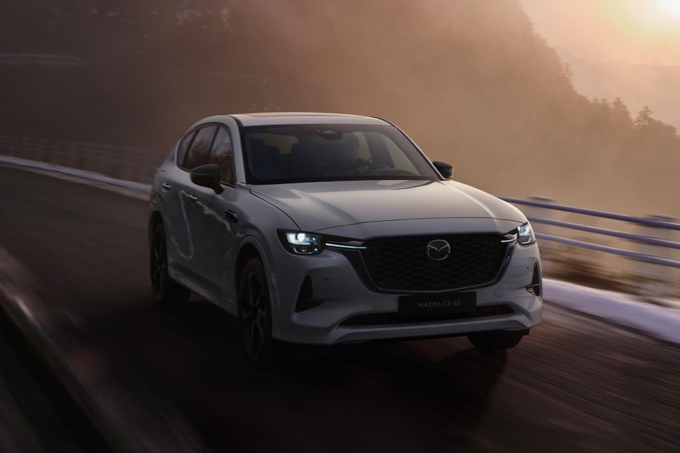 A light grey 2027 Mazda CX-60 SUV driving along a misty mountain road at sunrise, seen from the front three-quarter angle
