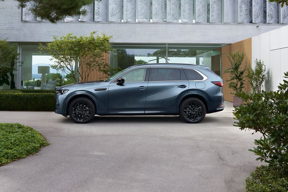 A blue-grey 2027 Mazda CX-80 SUV with black alloy wheels parked in profile in front of a modern glass building