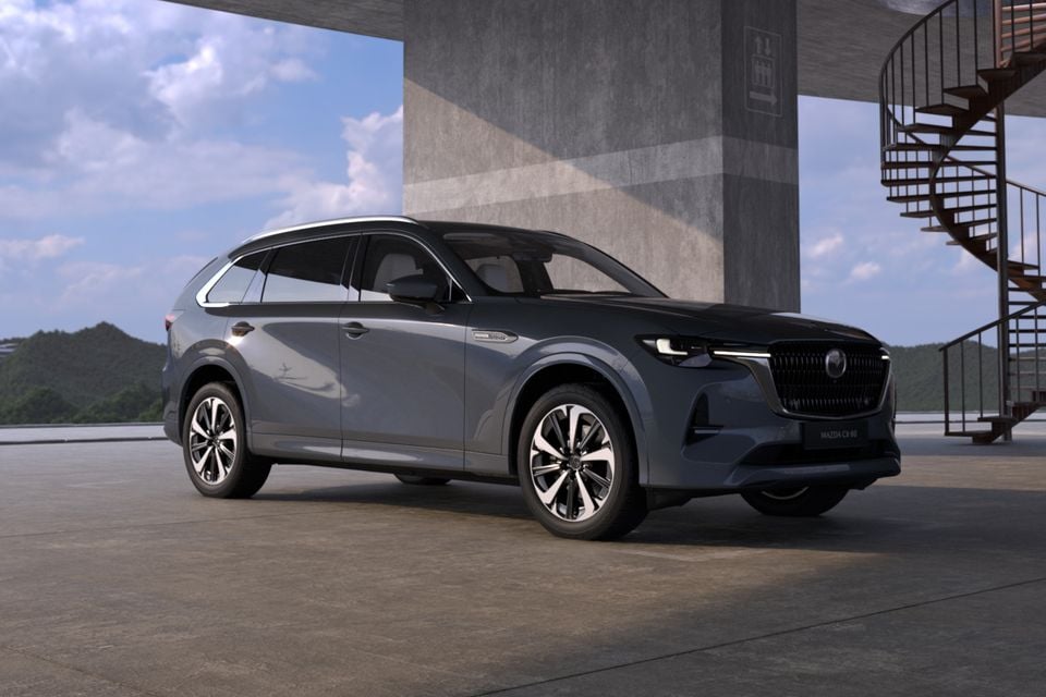 A grey 2027 Mazda CX-80 SUV parked beneath a concrete structure with a spiral staircase, seen from the front passenger side