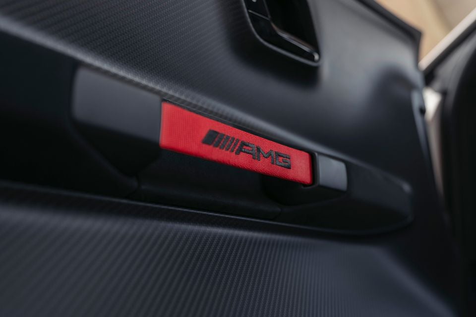 Close-up of a red AMG-branded door pull strap inside a Mercedes-AMG CLE, set against dark carbon-fibre-effect trim.