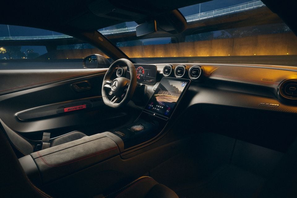 Interior of a Mercedes-AMG CLE showing the flat-bottomed steering wheel, large central touchscreen, carbon fibre trim, AMG-branded door sill, and sport seats with red stitching