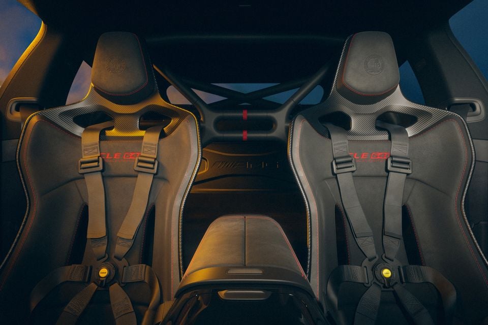Interior view of a Mercedes-AMG CLE 63 showing two carbon-fibre sport seats with harness-style seatbelts and red stitching accents, with a roll cage visible in the background