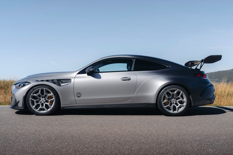 A silver Mercedes-AMG CLE coupe seen from the side, featuring large alloy wheels with orange brake calipers and a prominent rear wing, parked on a road with a rural backdrop.