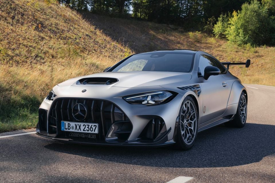 A grey Mercedes-AMG coupe seen from the front-side angle on a rural road, featuring a large front grille, hood scoop, rear wing, and aggressive aerodynamic bodywork.